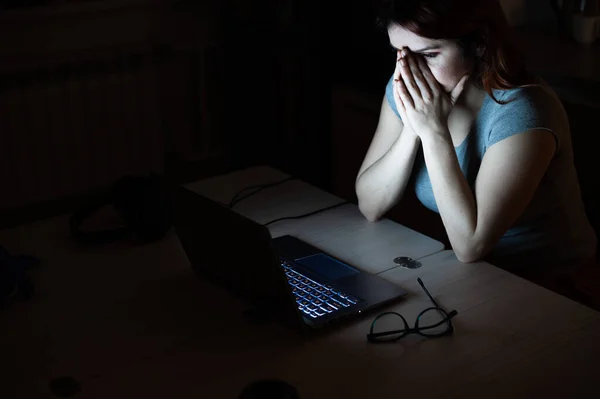 woman works overtime. A student studying at a laptop at night in the dark. The girl is worried about problems at work and holds her hands on her face.