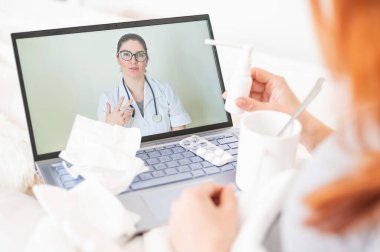 Bir kadın yatağa uzanır ve boğaz ağrısı için sprey tutar. Dost canlısı bir doktor hasta bir hastayla video bağlantısıyla konuşur. Laptop ekranında terapist. Karantina sırasında çevrimiçi tıbbi konsültasyon.