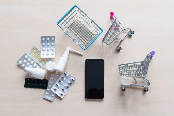 Online pharmacy concept. Buying medicine online over the phone. Delivery of pharmaceuticals. Mini trolley with different tablets, sprays, capsules and a smartphone with a black blank screen.