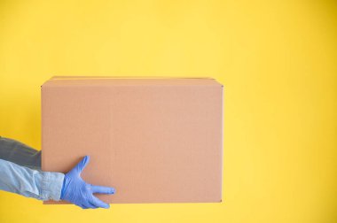 Eldivenli kadın elleri sarı arka planda küçük bir karton kutu tutuyor. Ev teslimatı yapan bir mağaza. Kot gömlekli bir kurye müşteriye bir karton çanta uzatır..