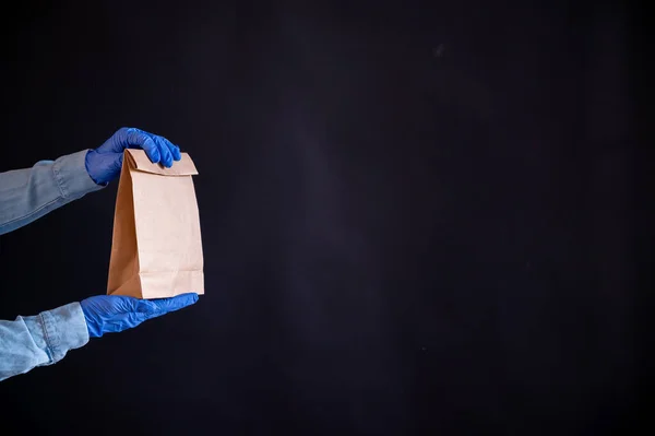 Las manos femeninas en un guante azul sostienen una bolsa de papel marrón sobre un fondo negro. Entrega de comida segura a su hogar. Un mensajero en una camisa de mezclilla sostiene una bolsa de cartón artesanal a un cliente . — Foto de Stock