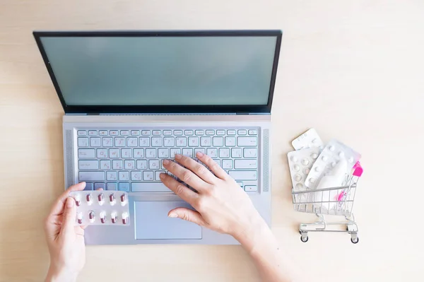 Online pharmacy. Closeup of female hands on a laptop and tablet keyboard in a mini shopping trolley with tablets. A woman chooses medicines on the Internet with home delivery.