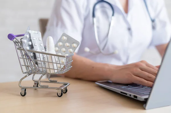 Female doctor working on a laptop. Mini shopping trolley full of pills. Online pharmacy concept. A female pharmacist is selling drugs over the Internet.