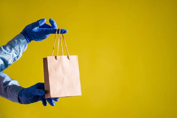 Female hands in gloves holds a small brown paper bag with handles on a yellow background. Safe food delivery to your home. A courier in a denim shirt holds out a craft cardboard bag to a customer. — Stock Photo, Image