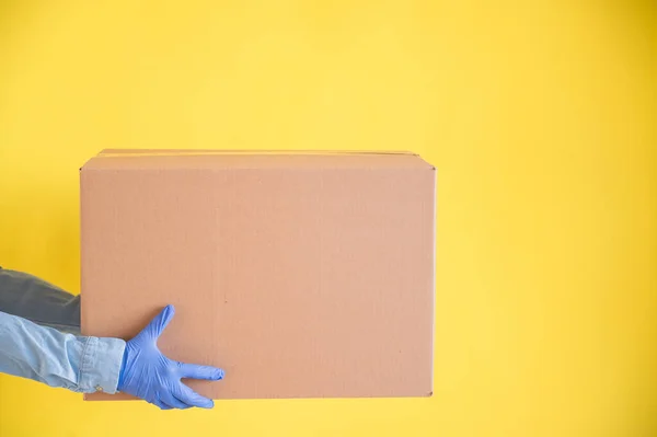 Close-up female hands in gloves holds a small cardboard box on a yellow background. Online store with home delivery. A courier in a denim shirt holds out a craft cardboard bag to a customer. — Stock Photo, Image