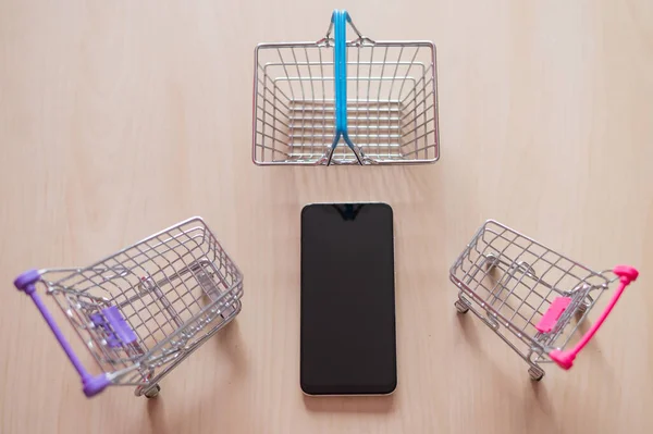 Κάτοψη σε κινητό τηλέφωνο δύο καροτσάκια και ένα καλάθι σε ξύλινο τραπέζι. Online αγορές σε ένα smartphone. Προεπισκοπήσεις. — Φωτογραφία Αρχείου