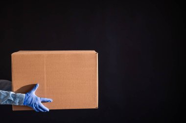 Eldivenli ve kot gömlekli kadın elleri. Teslimatçı karton kutuyu siyah arka planda müşteriye verir. Karantinada antimikrop koruması. Kırpılmış.