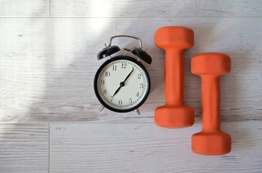 Bir çift turuncu dambıl ve çalar saatin üst görüntüsü. Sabahın köründe spor yapma fikri. Evde kendini eğitme zamanı..