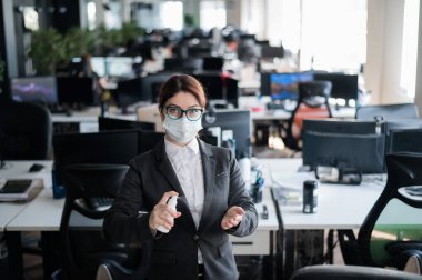 Tıbbi maske takmış kadın yönetici el antiseptiğine sprey sıkar. Takım elbiseli bir kadın dezenfekte için dezenfektan kullanıyor. Coronavirüs salgını sırasında ofis işi kavramı.