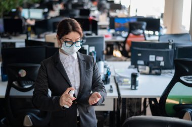 Tıbbi maske takmış kadın yönetici el antiseptiğine sprey sıkar. Takım elbiseli bir kadın dezenfekte için dezenfektan kullanıyor. Coronavirüs salgını sırasında ofis işi kavramı.