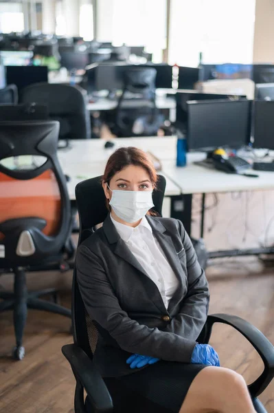 Female manager is sitting in a chair at the workplace. Business woman in mask and gloves at the desk in the office. Concept of work during a virus epidemic.