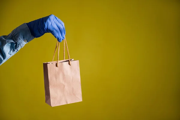 Female hands in gloves holds a small brown paper bag with handles on a yellow background. Safe food delivery to your home. A courier in a denim shirt holds out a craft cardboard bag to a customer. — Stock Photo, Image