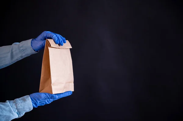 Female hands in a blue glove holds a brown paper bag on a black background. Safe food delivery to your home. A courier in a denim shirt holds out a craft cardboard bag to a customer. — Stock Photo, Image