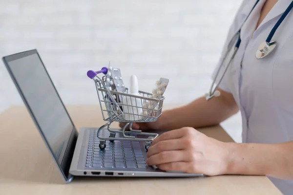 Felismerhetetlen női orvos a laptopon. Online gyógyszertár koncepció. Az arctalan gyógyszerész egy mini bevásárlókocsiba teszi a gyógyszert.. — Stock Fotó