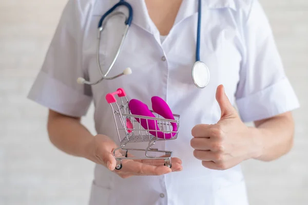 The faceless gynecologist recommends buying a clitoral vaginal vibrator to maintain womens health. The doctor holds a mini trolley and a masturbator for vivid orgasms. Gesture thumb.