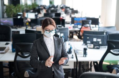 Tıbbi maske takmış kadın yönetici el antiseptiğine sprey sıkar. Takım elbiseli bir kadın dezenfekte için dezenfektan kullanıyor. Coronavirüs salgını sırasında ofis işi kavramı.