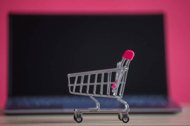 İnternetten alışveriş konsepti. Mini market arabası ve pembe arka planda dizüstü bilgisayar. Küçük araba ve kişisel bilgisayar.