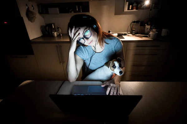Femme fatiguée dans des écouteurs et des lunettes étudiant sur un ordinateur portable. Une étudiante se prépare pour un examen dans la cuisine tard dans la soirée. Fille avec un chiot sur les genoux . — Photo