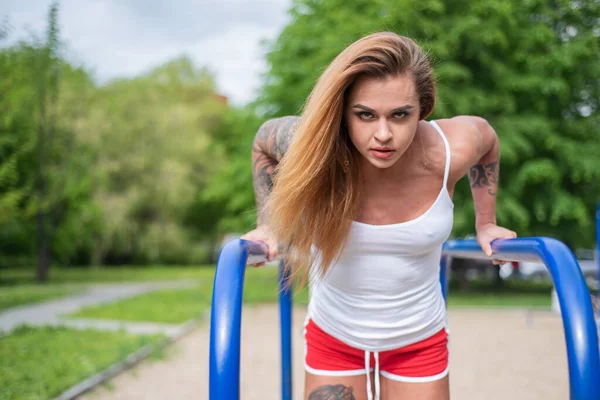 Donna atletica che fa esercizi tricipiti sulle sbarre irregolari sul parco giochi in una calda giornata estiva. Body builder femminile in pantaloncini e un breve top si allena all'aperto durante la quarantena. goffrato corpo muscolare . — Foto Stock