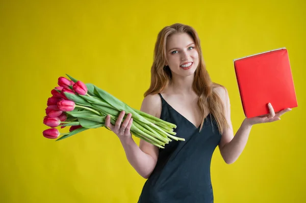 Žena s kyticí červených tulipánů na žlutém pozadí. Šťastná dívka v černých šatech drží náruč plnou květin a bonboniéru. Dárek na Valentýna. Nejromantičtější den v roce. — Stock fotografie