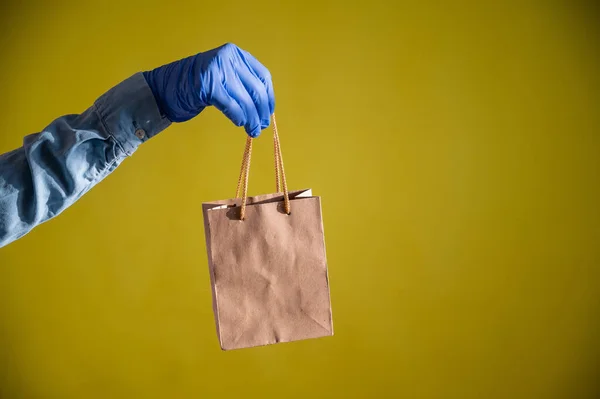 Female hands in gloves holds a small brown paper bag with handles on a yellow background. Safe food delivery to your home. A courier in a denim shirt holds out a craft cardboard bag to a customer.