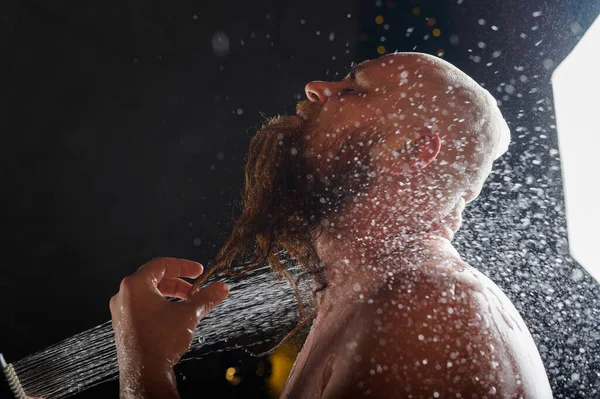 The bald guy takes a shower. A brutal man with a red beard is standing in the bathroom under a stream of water and washes. Spray scatter on a black background.