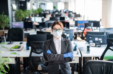 İş kadını ofiste kollarını kavuşturmuş duruyor. Tıbbi maske ve eldiven takan kadın yönetici. Bir koronavirüs salgınında çalışmak. Enfeksiyonun yayılmasını önleyecek yollar. İş yeri dezenfeksiyonu.