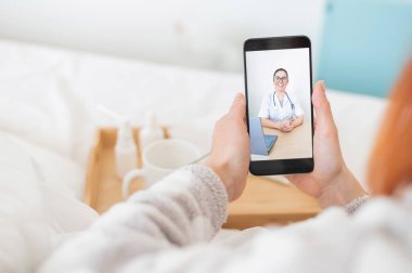 Mesafe tedavisi kavramı. Pijamalı, elinde akıllı telefon olan bir kadının dikiz görüntüsü. Bir grip hastası tıbbi video blogu izliyor..