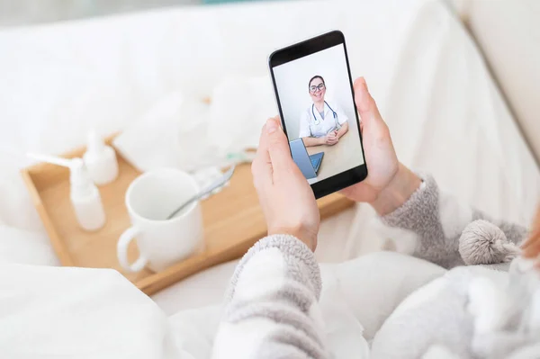 女人穿着睡衣躺在床上手里拿着智能手机一名流感患者正在看医疗视频博客。一位友善的医生通过电话提供在线咨询. — 图库照片