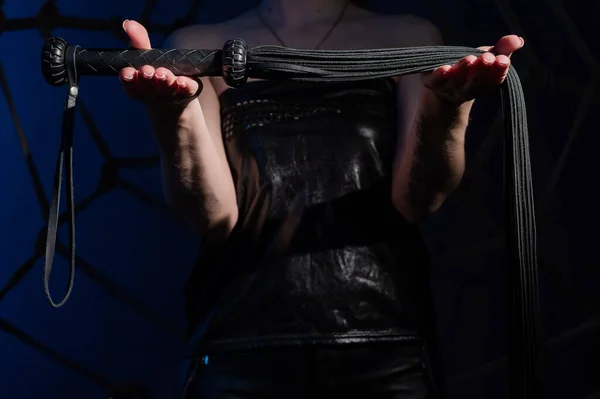 Black leather lash in the hands of an unrecognizable woman. Cropped photo of bdsms toys. Punishment and pleasure. A woman stands in the dark and holds a whip in her palms. — Stock Photo, Image