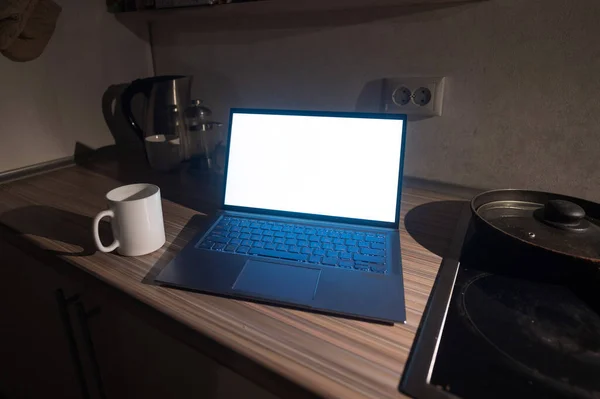 Un ordinateur portable de travail avec un écran blanc se tient sur la table de cuisine à côté de la cuisinière dans le noir. Le concept de télétravail. Quarantaine pendant le coronavirus . — Photo