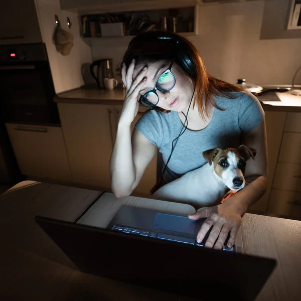 Femme fatiguée dans des écouteurs et des lunettes travaillant des heures supplémentaires sur un ordinateur portable. Femme pigiste travaillant à distance à la maison dans la cuisine tard dans la soirée. Fille avec un chiot sur les genoux . — Photo