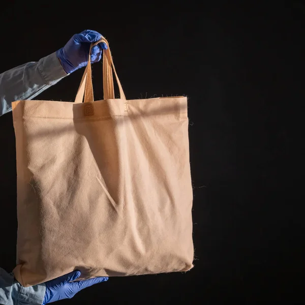 En påse bomullstyg på svart bakgrund. Kuriren håller en duk tygpåse utan plastförpackning på en svart bakgrund. Säker leverans till epidemin. Närbild av händer i handskar. — Stockfoto