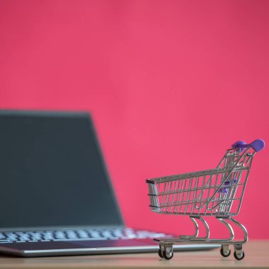 İnternetten alışveriş konsepti. Mini market arabası ve pembe arka planda dizüstü bilgisayar. Küçük araba ve kişisel bilgisayar.