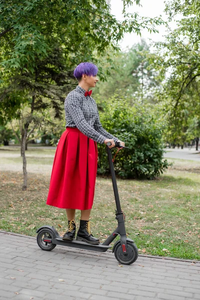 Egy fiatal nő, lila hajú lovagol egy elektromos robogó egy parkban. Stílusos lány egy kockás ing, egy hosszú vörös szoknya és egy csokornyakkendő a lovaglás a város körül egy modern eszköz. Csípő. — Stock Fotó