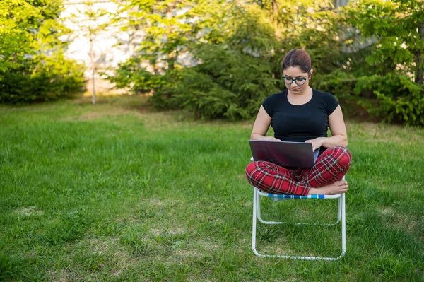 Uma mulher com óculos senta-se em uma cadeira dobrável e digita em um laptop. Freelancer feminino trabalha remotamente no jardim da casa de campo. Trabalho ao ar livre durante a quarentena . — Fotografia de Stock