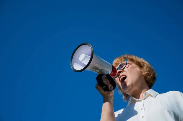 Eine ältere Frau mit Brille ruft im Freien in ein Megafon. Eine genervte Rentnerin stellt ihre Forderungen mit Hilfe eines dreifachen Verstärkungssignals vor blauem Hintergrund. Lautsprecher in Frauenhand. — Stockfoto