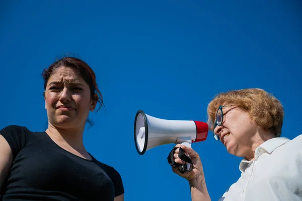 Конфликт поколений. Эмоциональная пожилая женщина кричит на свою дочь в мегафоне. Пожилая мать ругается на женщину средних лет на громкоговорителе на синем фоне . — стоковое фото