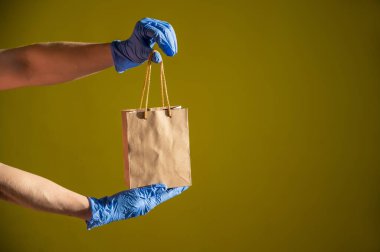 Tıbbi lateks eldivenli kadın kurye küçük bir kese kağıdı tutuyor. Koronavirüs yayılırken güvenli bir teslimat. Çevre dostu ambalaj. İnternetten alışveriş.