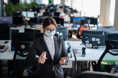 Tıbbi maske takmış kadın yönetici el antiseptiğine sprey sıkar. Takım elbiseli bir kadın dezenfekte için dezenfektan kullanıyor. Coronavirüs salgını sırasında ofis işi kavramı.