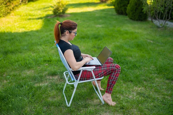 Uma mulher com óculos senta-se em uma cadeira dobrável e digita em um laptop. Freelancer feminino trabalha remotamente no jardim da casa de campo durante a quarentena. O trabalho é um prazer. . — Fotografia de Stock