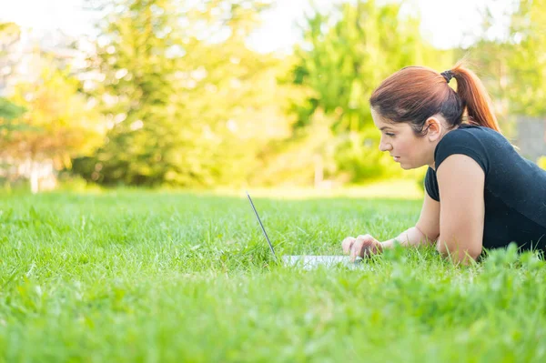 Uma mulher deita-se de barriga em um parque e digita em um laptop. Freelancer feminino trabalha remotamente ao ar livre. Menina está estudando em um computador no gramado . — Fotografia de Stock