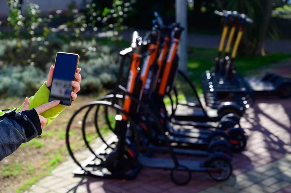 La chica sin rostro utiliza la aplicación móvil para pagar y desbloquear el scooter eléctrico de alquiler. La mano femenina de cerca con un teléfono. Mujer en transporte ecológico para moverse por la ciudad . — Foto de Stock