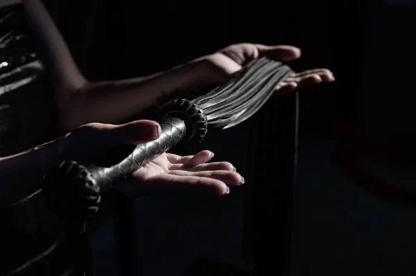 Close-up chicote de couro preto em mãos femininas. Monocromático foto recortada de brinquedos bdsms. Uma mulher fica no escuro e segura um chicote nas palmas das mãos . — Fotografia de Stock