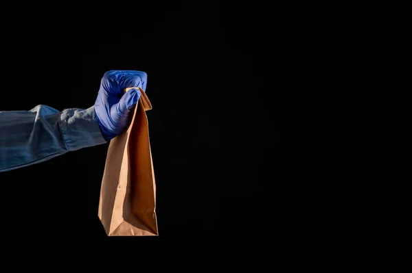 Kvinnlig hand i en blå handske håller en brun papperspåse på en svart bakgrund. Säker mat leverans till ditt hem. En kvinna i jeansskjorta håller fram en pappsäck till en kund. — Stockfoto