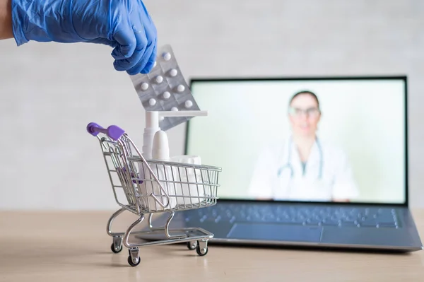 Egy barátságos orvos online konzultációt folytat egy laptopon. Egy kesztyűs gyógyszerész gyógyszereket tesz egy mini bevásárlókocsiba. A távorvoslás fogalma. Rendeljen online gyógyszereket. — Stock Fotó