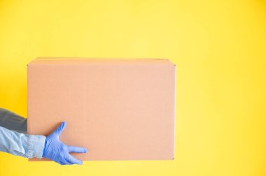 Eldivenli kadın elleri sarı arka planda küçük bir karton kutu tutuyor. Ev teslimatı yapan bir mağaza. Kot gömlekli bir kurye müşteriye bir karton çanta uzatır..