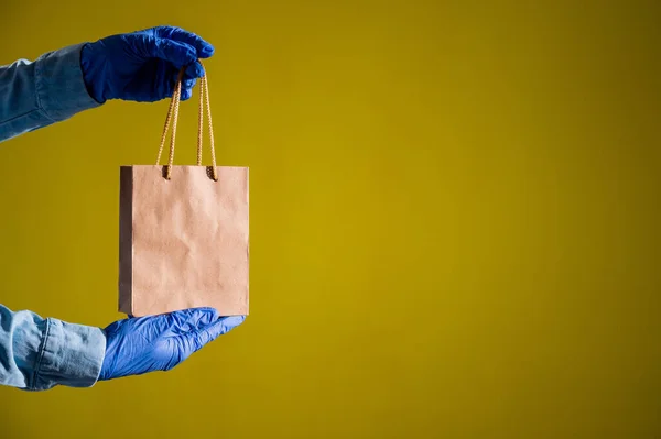 Female courier hand in medical latex gloves holds a small paper bag. Safe delivery during the spread of coronavirus. Ecofriendly packaging. Online shopping. — Stock Photo, Image