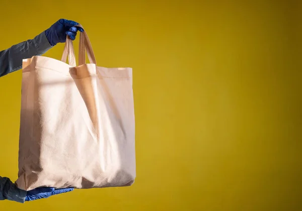 Enviando eco-friendly saco de algodão reutilizável em um fundo amarelo. O mensageiro segura um saco de tecido de lona sem embalagem de plástico em um fundo amarelo. Entrega segura à epidemia . — Fotografia de Stock