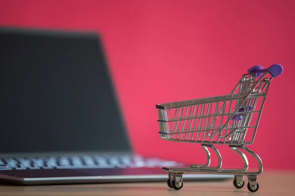 Concepto de compras online. Mini carrito de comestibles y portátil sobre un fondo rosa. Carro pequeño y ordenador personal . —  Fotos de Stock